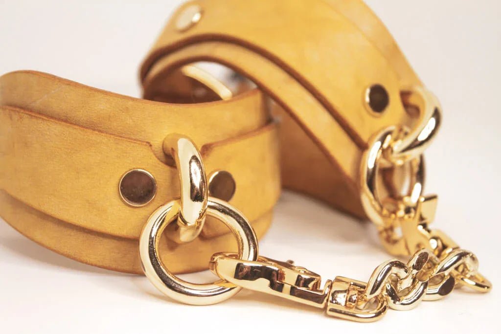 Golden Hour fine leather handmade BDSM cuffs connected by a gold-tone chain on a plain backdrop.