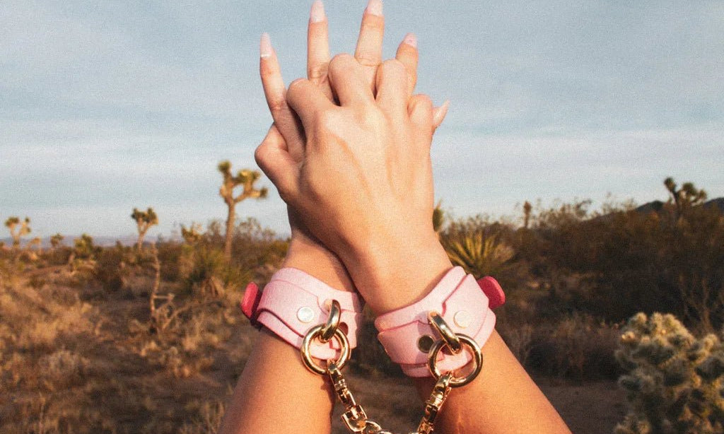 Close-up of hands wearing pink leather wrist cuffs connected by a gold chain in a desert landscape