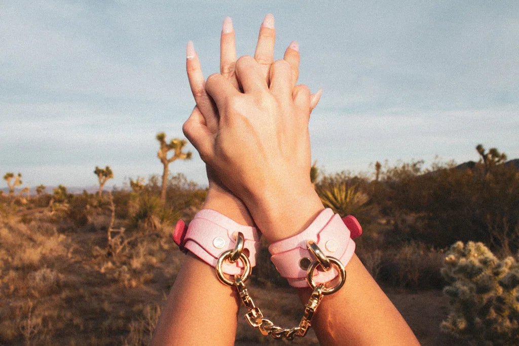 Close-up of hands wearing pink leather wrist cuffs connected by a gold chain in a desert landscape