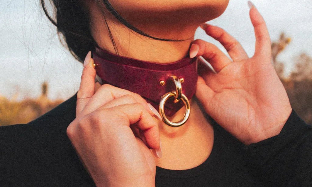 Close-up of a person wearing a burgundy leather day collar with a large gold ring, adjusting it with their hands outdoors
