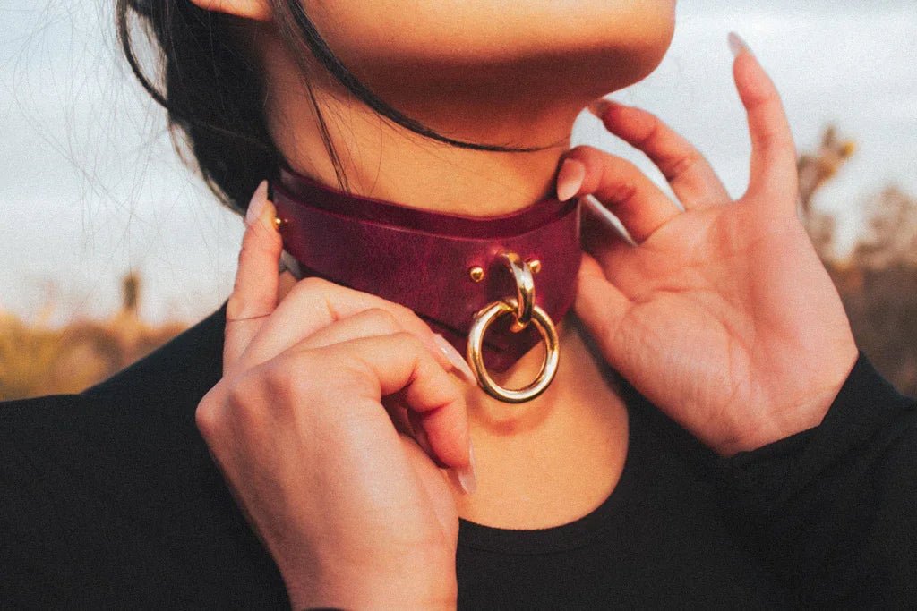 Close-up of a person wearing a burgundy leather day collar with a large gold ring, adjusting it with their hands outdoors
