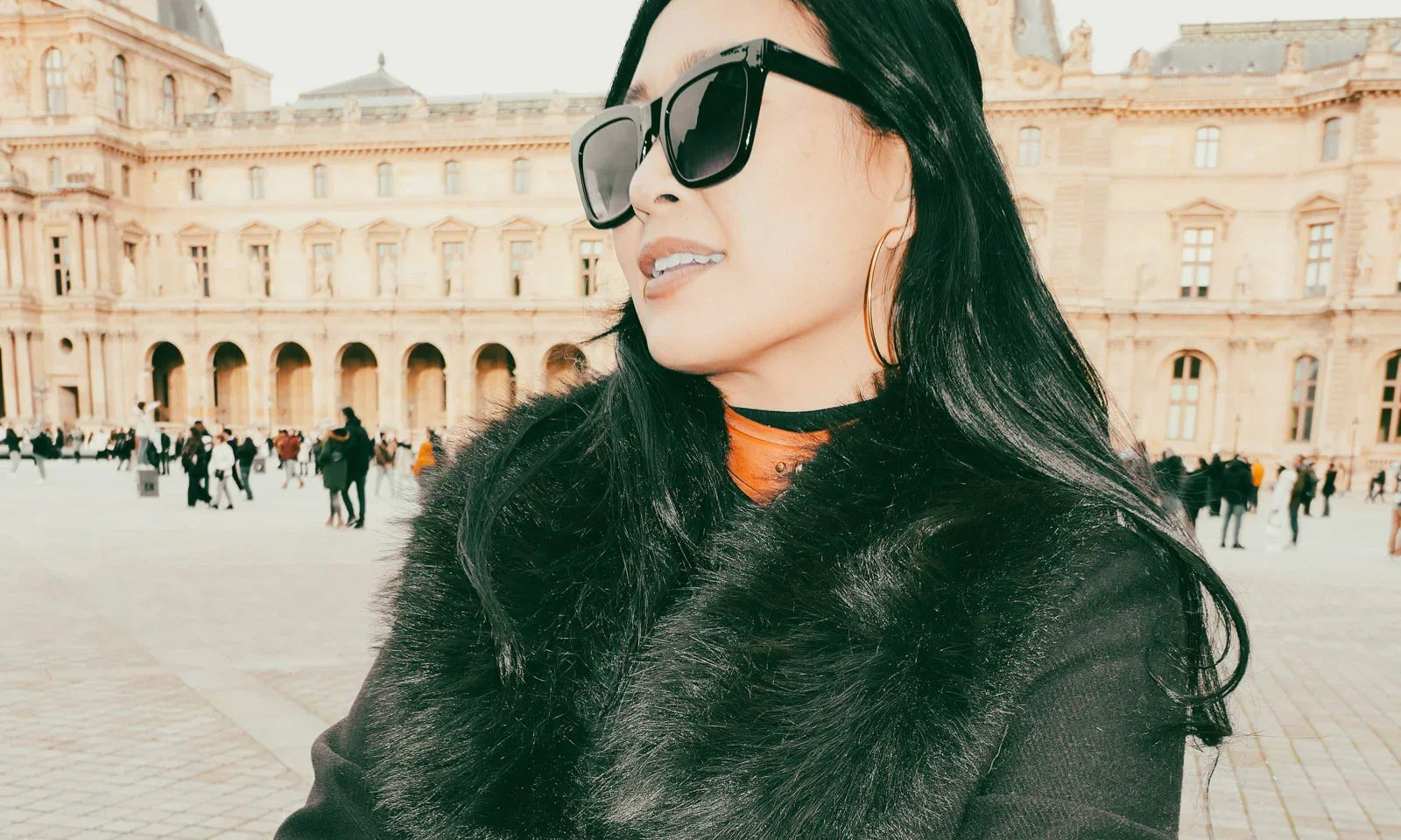 Woman wearing black leather day collar and black fur coat with large sunglasses in front of historic building