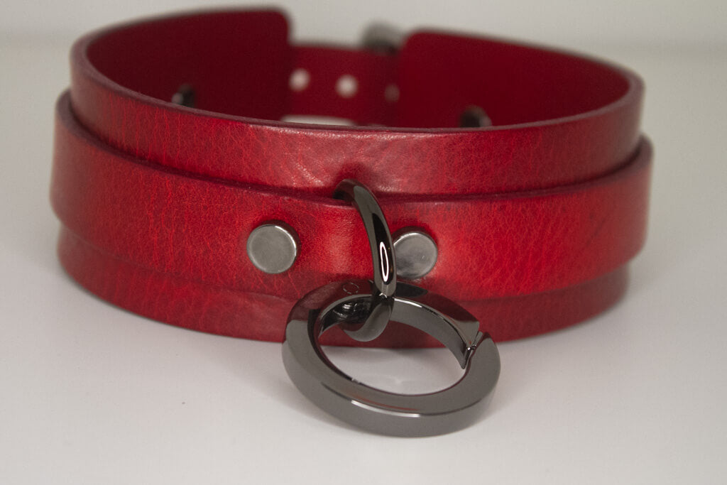 Close-up view of the 'Blood Moon' red leather BDSM collar resting on a flat surface, focusing on the black O-ring and the collar's texture and craftsmanship.