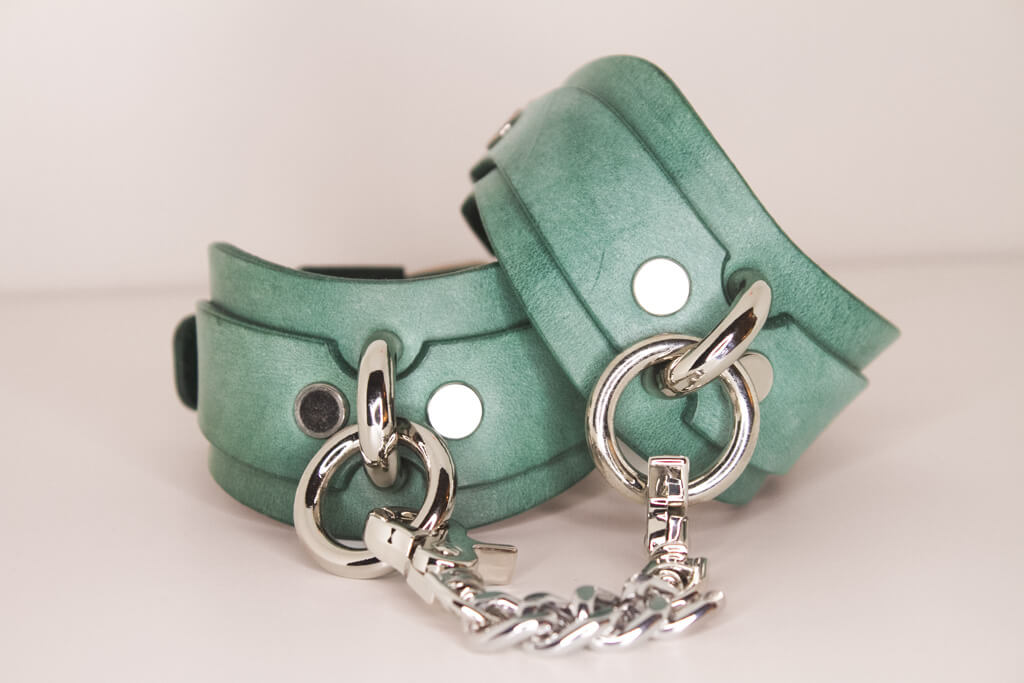 Elegantly crafted teal Italian leather BDSM cuffs with silver hardware, set against a white background.