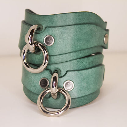 Stack of handmade Italian leather kinky cuffs in teal with silver O-rings, luxury BDSM accessory.