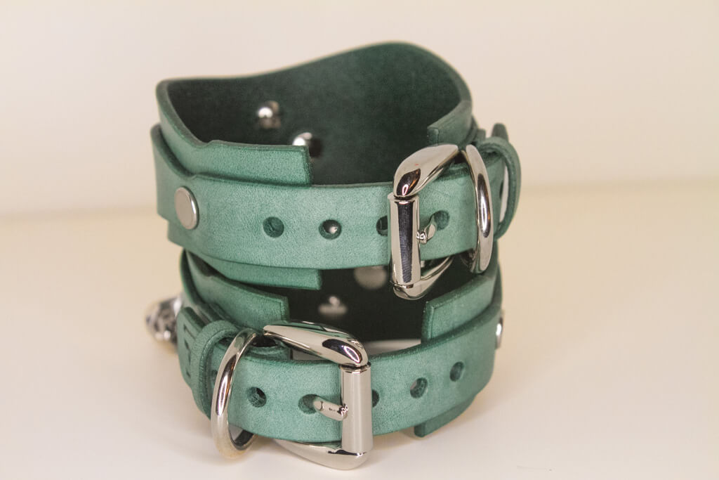 Back detail of two handmade teal Italian leather cuffs connected by a silver chain, showcasing luxury and kink.