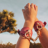 Hands wearing red leather wrist cuffs with gold chain outdoors near a tree sky background