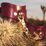 Wrist wearing Bordeaux leather BDSM cuffs with gold-tone hardware, desert scenery backdrop.
