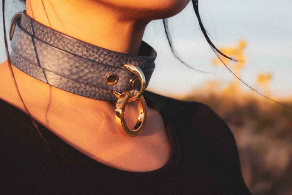 Lifestyle shot of a person wearing a denim leather BDSM collar, showcasing its fit and the gold-tone O-ring detail, set against a natural outdoor backdrop.