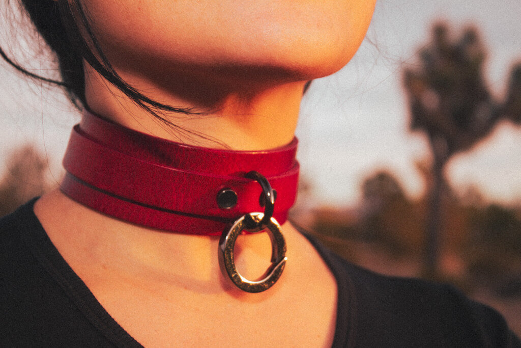 Front-facing lifestyle image of an individual wearing the 'Blood Moon' red leather BDSM collar, with the black O-ring detail highlighted against a blurred outdoor setting.