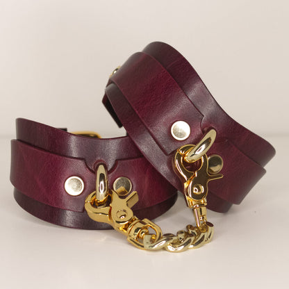 Bordeaux leather BDSM cuffs with gold-tone chain and clasps, set against a neutral background.