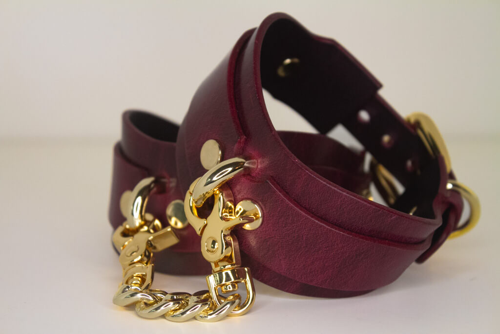 Stacked Bordeaux leather BDSM cuffs with gold-tone buckle closures, gold chains and rivet details.