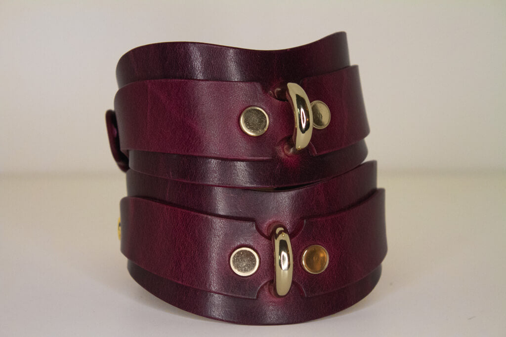 Stacked Bordeaux leather BDSM cuffs with gold-tone buckle closures and rivet details.