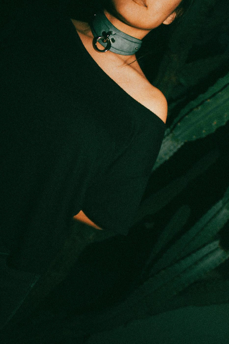 Person wearing a gray custom leather day collar with a metal ring, black off-shoulder top, dark background