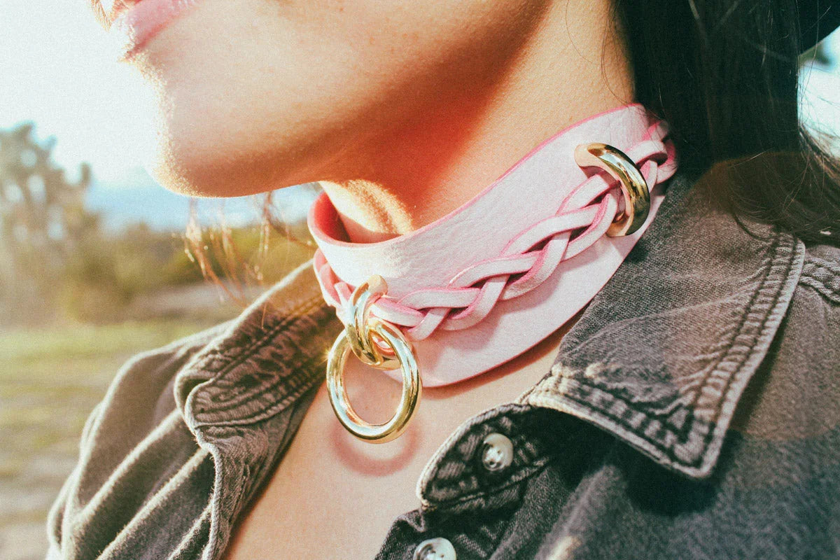 Close-up of a person wearing a pink braided leather day collar with gold rings, paired with a denim jacket