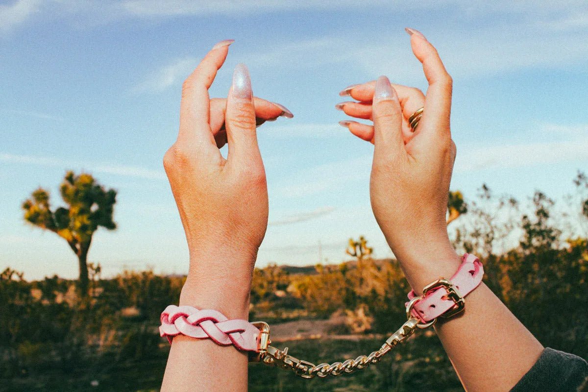 Hands wearing pink leather wrist cuffs connected by a gold chain against a desert background with blue sky