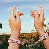 Pair of hands wearing pink leather wrist cuffs connected by a gold chain against a desert background under clear blue sky