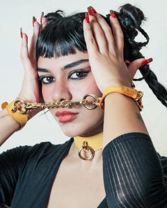 Woman wearing yellow leather day collar and matching leather wrist cuffs connected by a gold chain