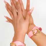 Wrists wearing pink leather wrist cuffs with gold rings on a neutral background, showcasing leather day collar style