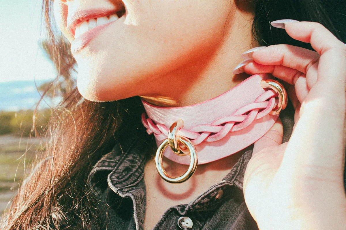 Close-up of a smiling woman wearing a pink leather day collar with braided detail and gold ring outdoors