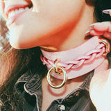 Close-up of a smiling woman wearing a pink leather day collar with braided detail and gold ring outdoors