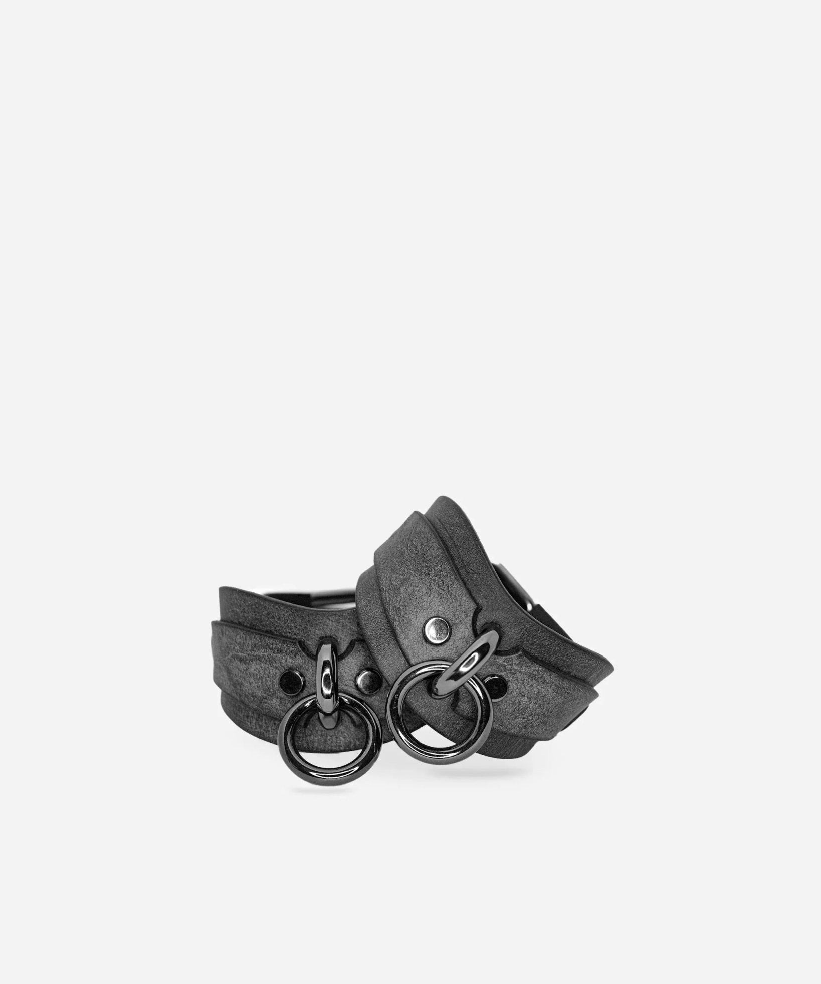Pair of dark leather wrist cuffs with metal rings, black hardware on plain background