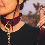 Woman wearing a burgundy leather day collar with gold chain detail holding matching leather wrist cuffs