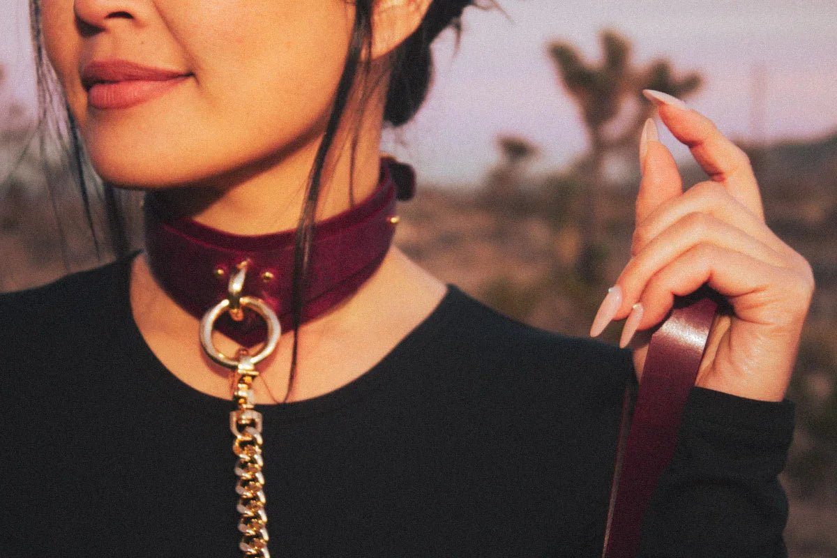 Woman wearing a burgundy leather day collar with gold chain detail holding matching leather wrist cuffs