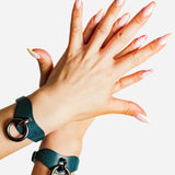 Hands wearing dark teal leather wrist cuffs with metal rings, showcasing custom leather cuffs and wrist accessories
