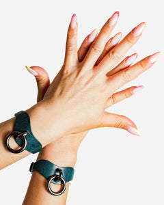 Hands wearing dark teal leather wrist cuffs with metal rings, showcasing custom leather cuffs and wrist accessories