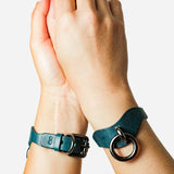 Close-up of hands wearing blue leather wrist cuffs with black metal buckles and rings on white background