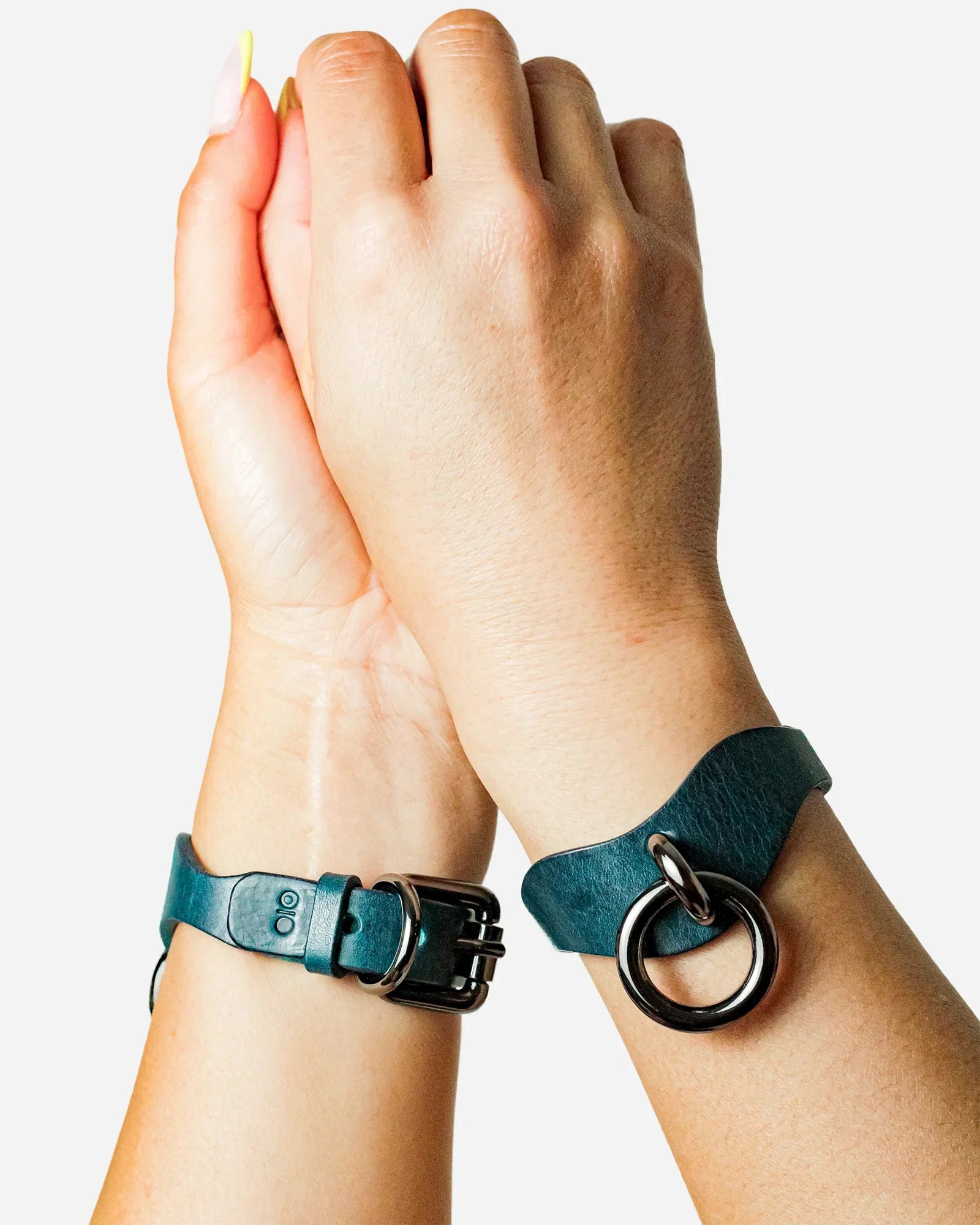 Close-up of hands wearing blue leather wrist cuffs with black metal buckles and rings on white background
