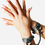 Hands wearing grey leather wrist cuffs connected by black metal chain against white background