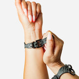 Person adjusting black leather wrist cuffs with metal rings and buckle on light skin background