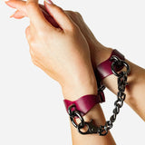 Hands wearing burgundy leather wrist cuffs connected by black metal chain on white background
