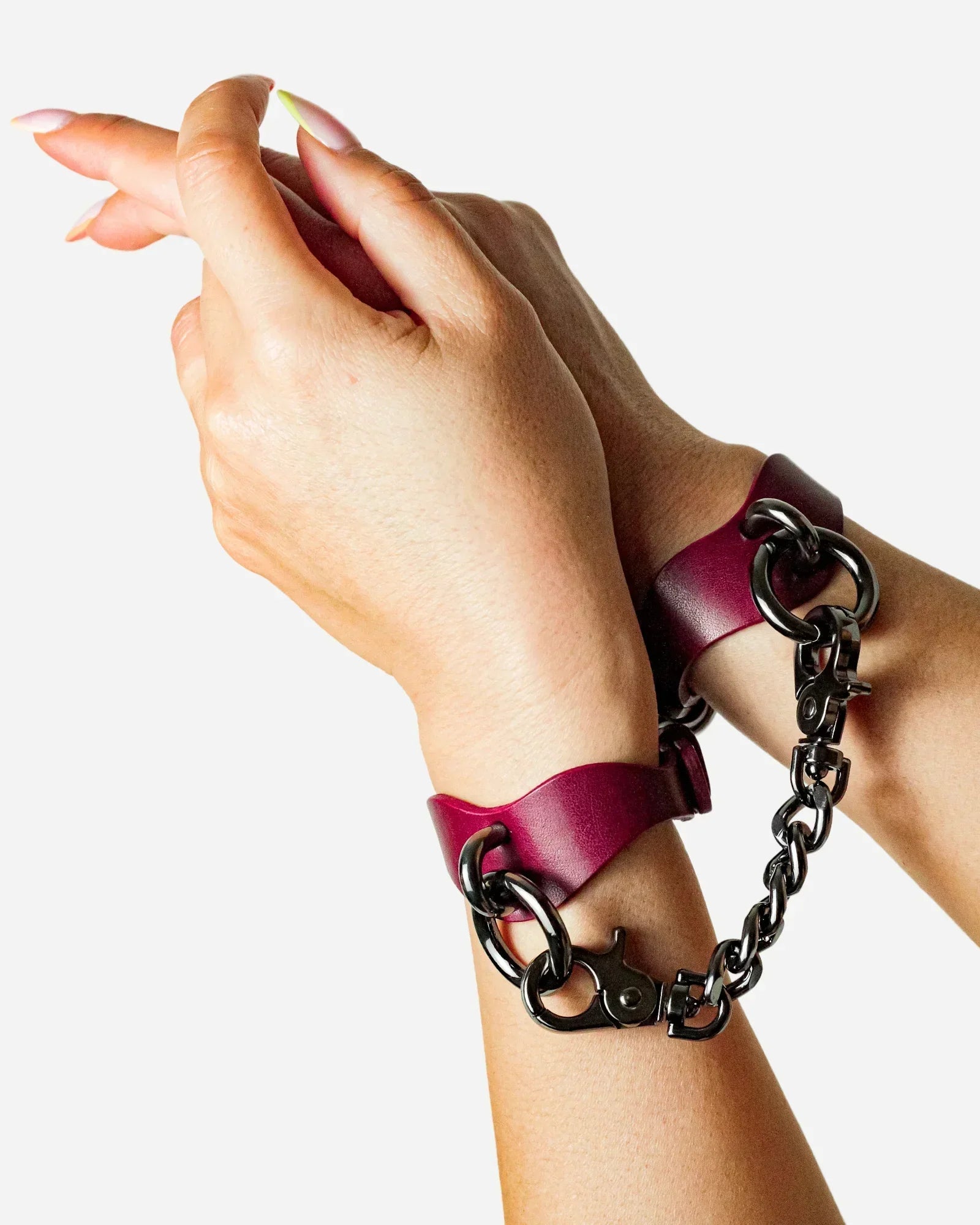 Hands wearing burgundy leather wrist cuffs connected by black metal chain on white background