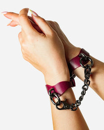 Hands wearing burgundy leather wrist cuffs connected by black metal chain on white background