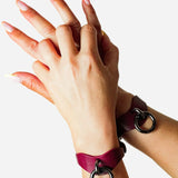 Hands wearing Bordeaux leather wrist cuffs with black metal rings against white background