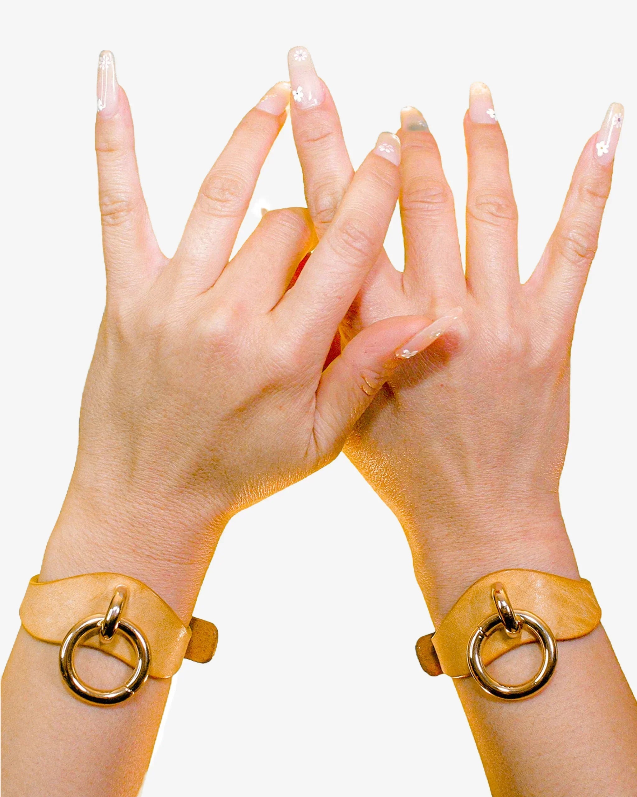 Hands wearing tan leather wrist cuffs with large gold rings against white background, showcasing leather cuffs