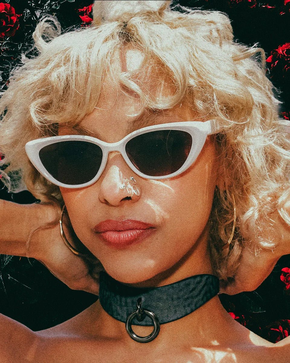 Close-up of woman wearing black leather day collar with ring, white cat-eye sunglasses, and gold hoop earrings