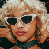 Close-up of woman wearing black leather day collar with ring, white cat-eye sunglasses, and gold hoop earrings