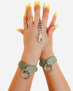 Hands wearing green leather wrist cuffs with metal rings and chain detail, styled with yellow nails