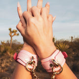 Hands clasped with pink leather wrist cuffs connected by gold chain in outdoor desert setting