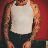 Tattooed man wearing a white tank top, red leather day collar, and black leather wrist cuffs against a red background