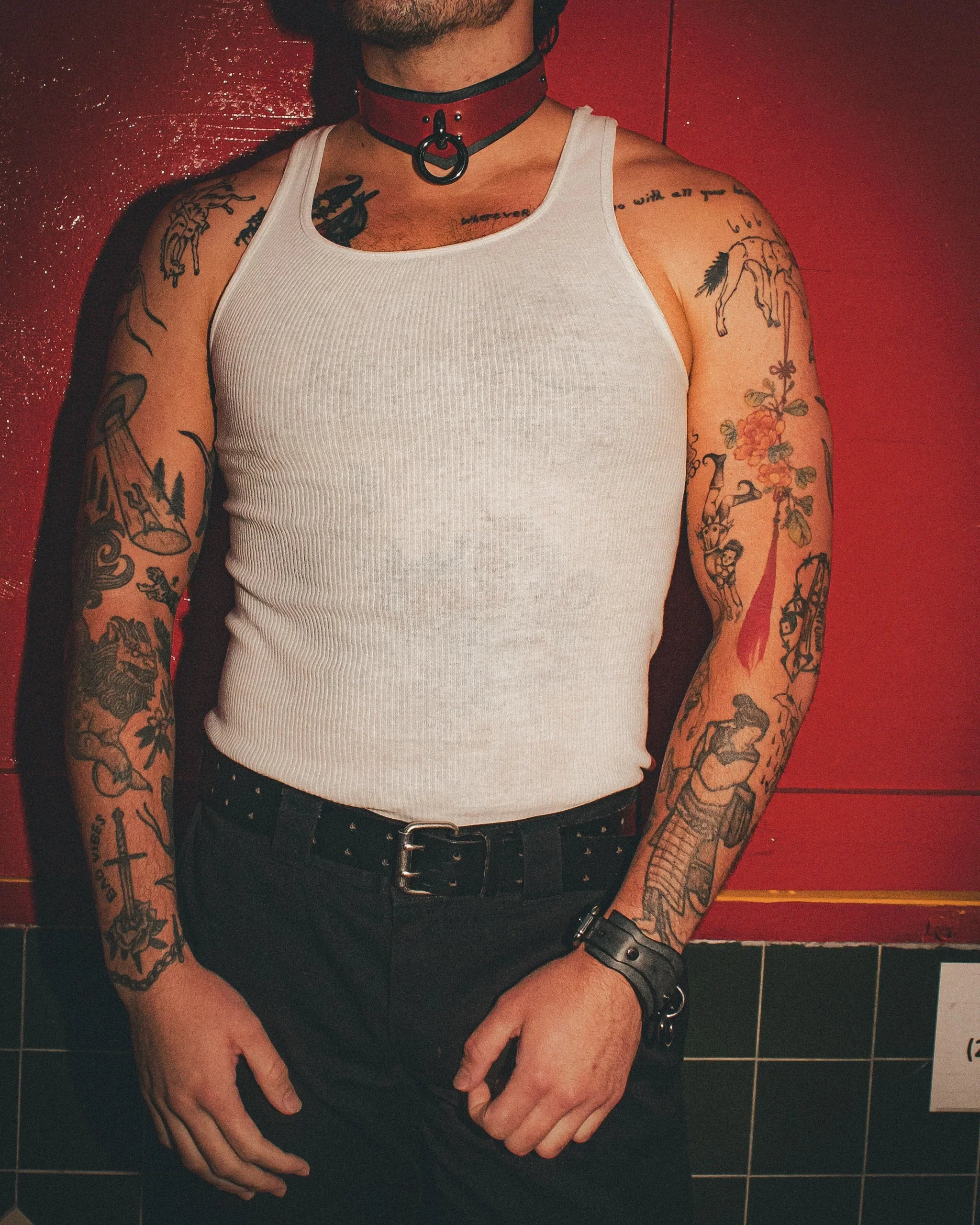 Tattooed man wearing a white tank top, red leather day collar, and black leather wrist cuffs against a red background