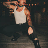 Man with tattoos wearing a white tank top and black pants sitting on a stone floor with string lights in the background. he's wearing a blue and black collar.