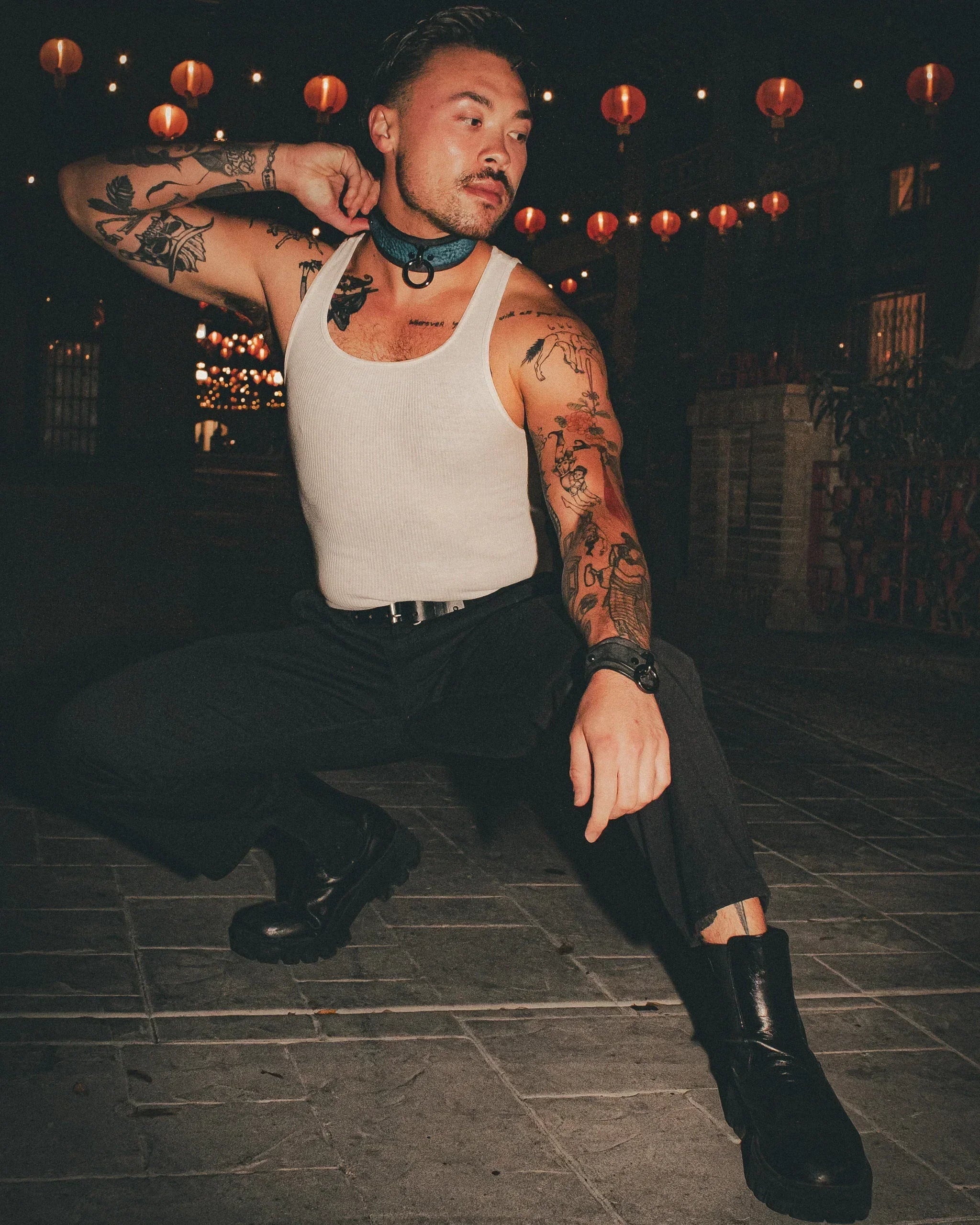 Man with tattoos wearing a white tank top and black pants sitting on a stone floor with string lights in the background. he's wearing a blue and black collar.