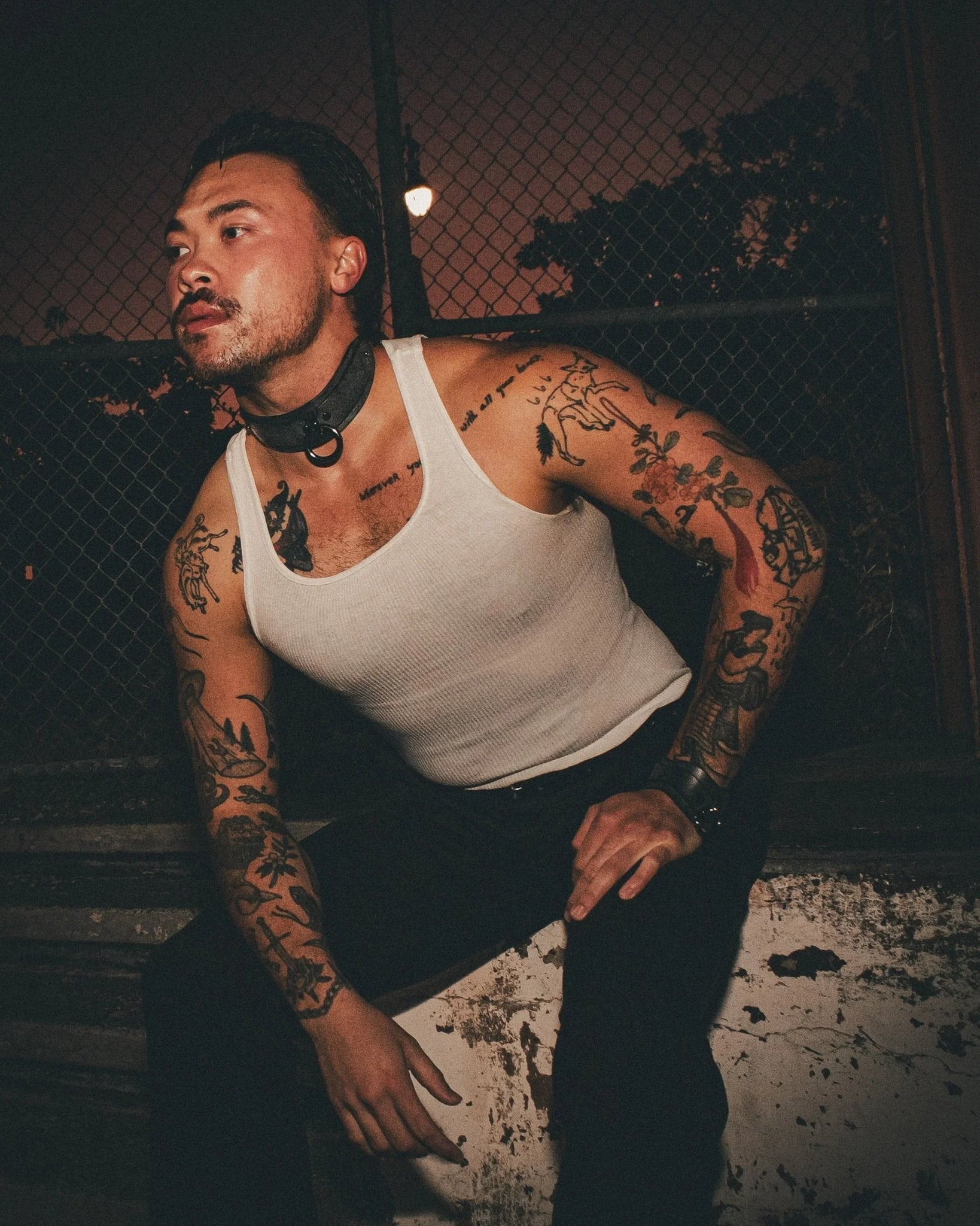 Tattooed man wearing white tank top with black leather day collar and leather wrist cuffs sitting by chain-link fence at night